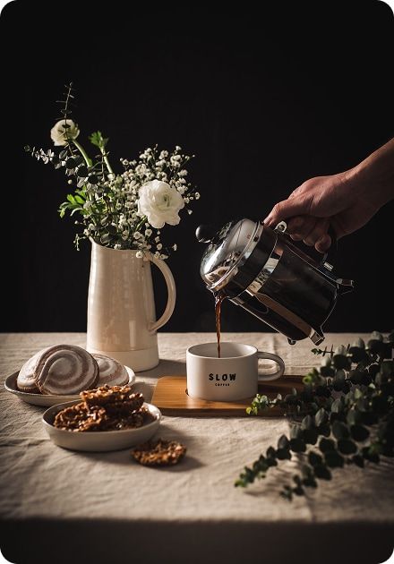 A hand pours coffee from a French press into a white mug labeled "SLOW COFFEE," next to a vase of white flowers and plates of pastries.