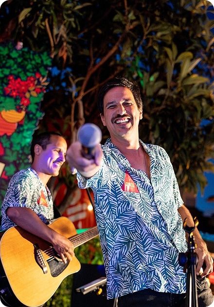Dois músicos atuam ao ar livre à noite; o homem em primeiro plano sorri e estende um microfone em direção à câmara, vestindo uma camisa estampada azul e branca, enquanto o músico ao fundo toca guitarra acústica e também usa camisa semelhante, com árvores e decoração colorida atrás deles.